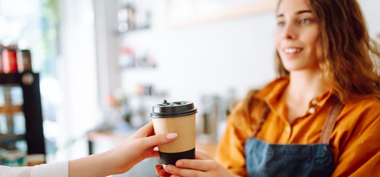 Jovem mulher atendendo cliente em cafeteria, segurando um copo de café para levar. O ambiente é acolhedor e descontraído.