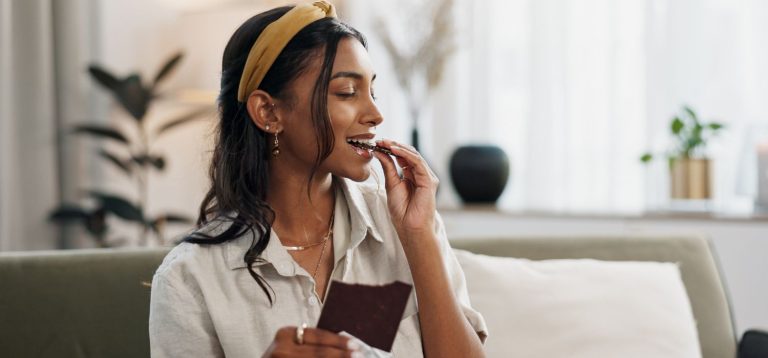 Imagem de uma jovem mulher comendo um chocolate enquanto relaxa em um sofá, transmitindo um momento de prazer e conforto.