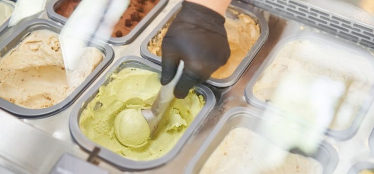 Imagem de um sorvete sendo servido em uma geladeira com vários sabores diferentes. Destaque para uma esfera de sorvete verde em destaque.