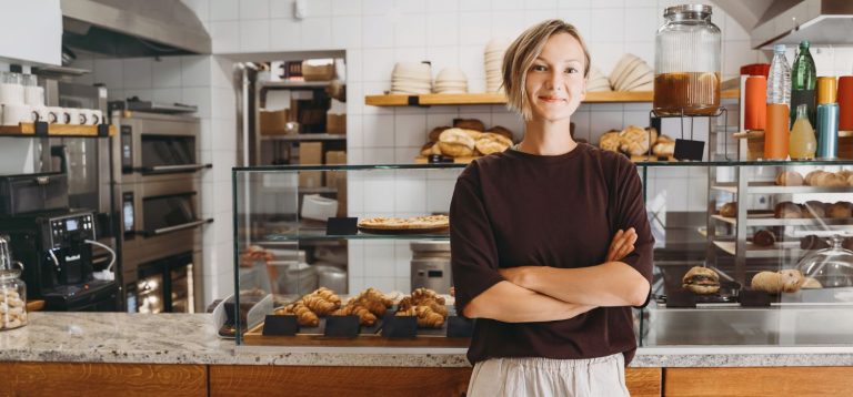 Mulher sorridente em uma padaria compacta e moderna com pães frescos expostos. Ambiente iluminado, transmitindo uma sensação acolhedora e agradável.
