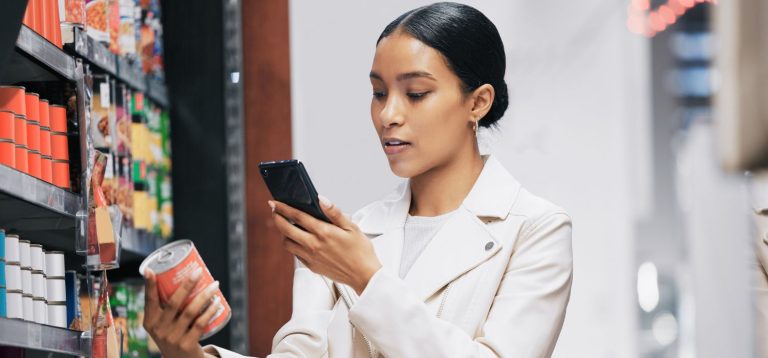 Mulher usando smartphone enquanto verifica produtos em prateleira de supermercado, mostrando hábitos de compra modernos e tecnologia no comércio.
