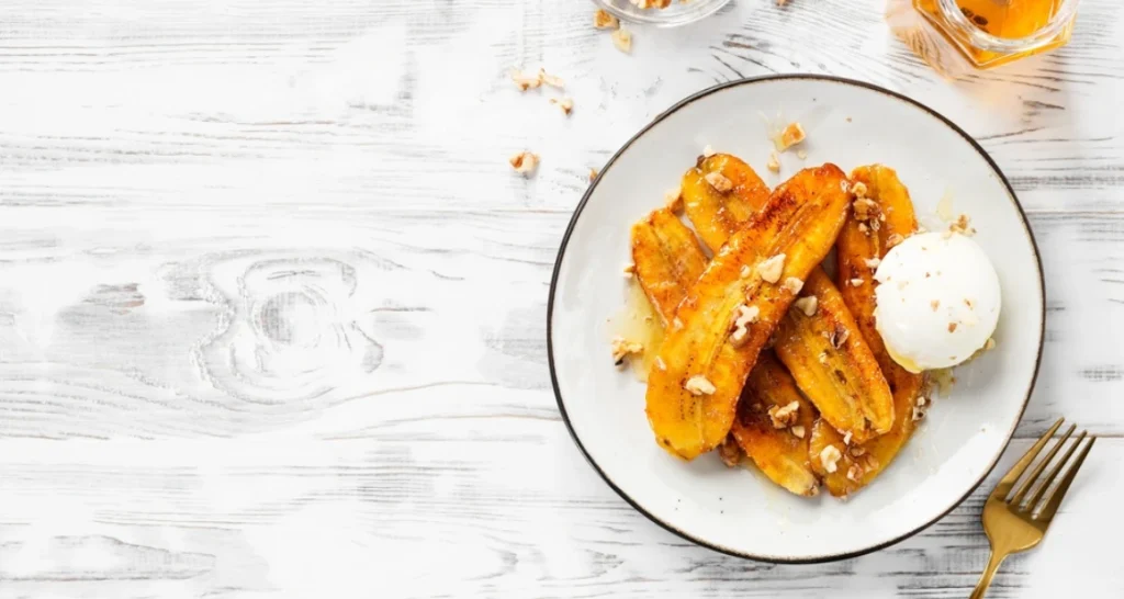 Banana caramelizada com sorvete de creme e castanhas, servida em prato branco sobre mesa de madeira clara.