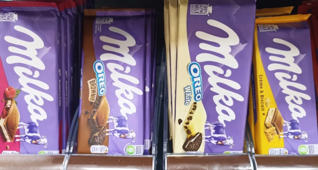Variedade de barras de chocolate Milka, incluindo sabores com Oreo, biscoito, White, brownie e creme com biscoito, em prateleira de supermercado.