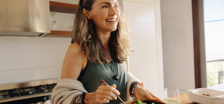 Mulher sorridente preparando uma refeição na cozinha, com utensílios de cozinha ao redor e um ambiente bem iluminado. A imagem reflete o tema "comida como terapia".