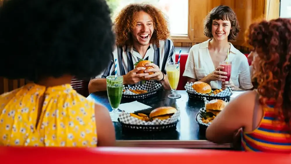 Grupo de mulheres sorridentes comendo hambúrgueres e tomando bebidas coloridas em uma confraternização alegre.