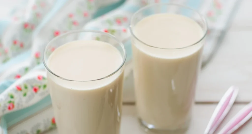 Dois copos de leite colocados sobre uma mesa de madeira clara, com uma toalha com detalhes de cereja ao fundo.