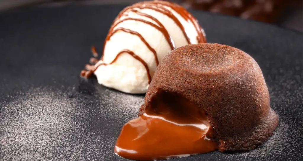 Sobremesa de petit gâteau de chocolate com sorvete de baunilha, decorado com calda de chocolate, servido em prato preto.