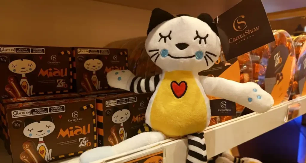 Gato de pelúcia com corpo amarelo, rosto branco, coração vermelho no peito, e detalhes pretos e brancos, posicionado em uma prateleira de loja da Cacau Show, com caixas de chocolates ao fundo.