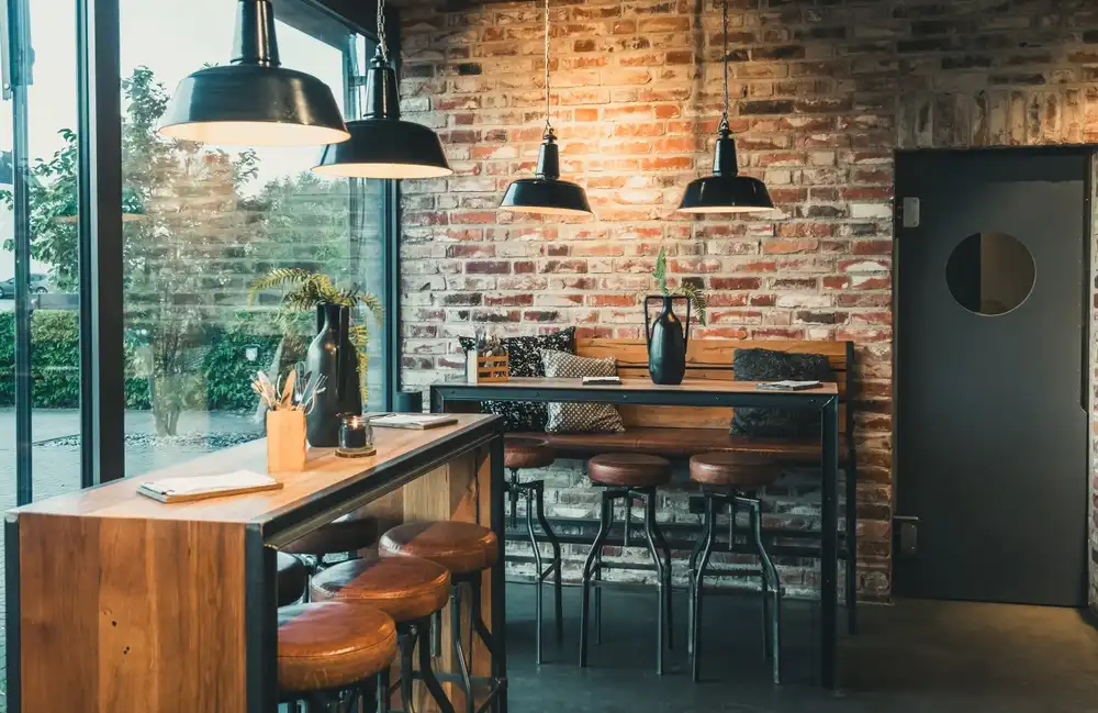 Interior de uma cafeteria moderna com parede de tijolos expostos, iluminação pendente, mesas de madeira, cadeiras de couro e uma grande janela de vidro.