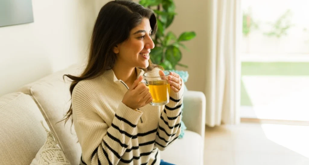 Mulher sorridente tomando chá de ervas em um ambiente confortável e bem iluminado.
