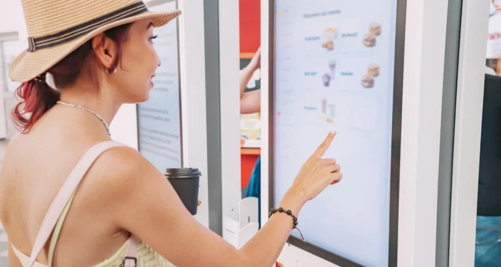 Mulher usando chapéu de palha interagindo com um quiosque digital em um shopping, consultando um menu de fast-food ou bebidas, na procura por opções rápidas e práticas.