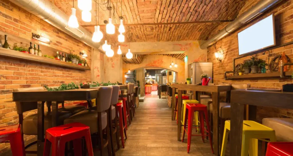 Interior de restaurante com decoração rústica, paredes de tijolos expostos, mesas de madeira e assentos coloridos, iluminação aconchegante e ambiente convidativo.