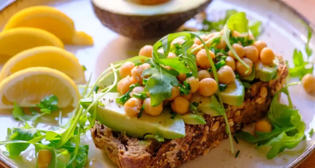 Torrada de pão integral com abacate, grão-de-bico, tomate e microverdes, acompanhada de fatias de limão, ideal para uma alimentação saudável e nutritiva.