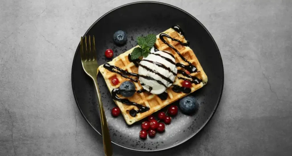 Waffle com sorvete de creme, mirtilos, framboesas, uvas e geleia de frutas, decorados com hortelã e calda de chocolate, servido em prato preto com garfo dourado.