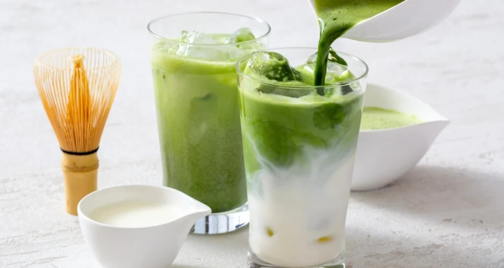 Imagem de drinks de matcha com leite, acompanhados de utensílios de cozinha como batedor de bambu e tigelas, sobre mesa branca de estilo minimalista.
