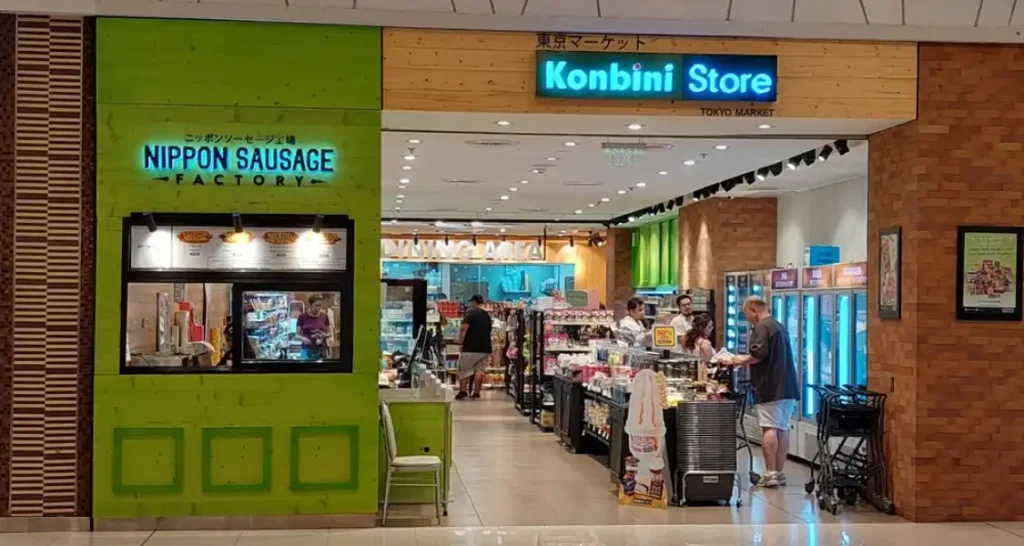 Entrada de loja no shopping Tokyo Market com destaque para o comércio Nippon Sausage Factory e a loja Konbini Store, com consumidores fazendo compras.