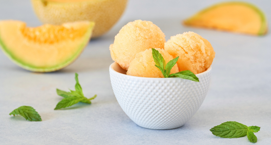 A imagem mostra uma taça branca com três bolas de sorvete vegano de melão, decoradas com folhas frescas de hortelã. Ao fundo, há fatias e um melão cortado, compondo uma estética fresca, natural e convidativa. O cenário é claro e minimalista, destacando a pureza e a simplicidade do produto.