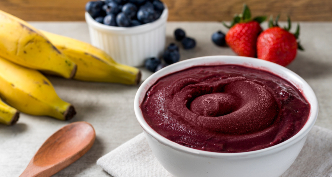 A imagem mostra uma tigela com um sorvete vegano de açaí cremoso, acompanhado por frutas naturais como bananas, morangos e blueberries ao fundo. A apresentação é simples, mas muito atraente e saudável, reforçando a ideia de um produto natural e nutritivo.