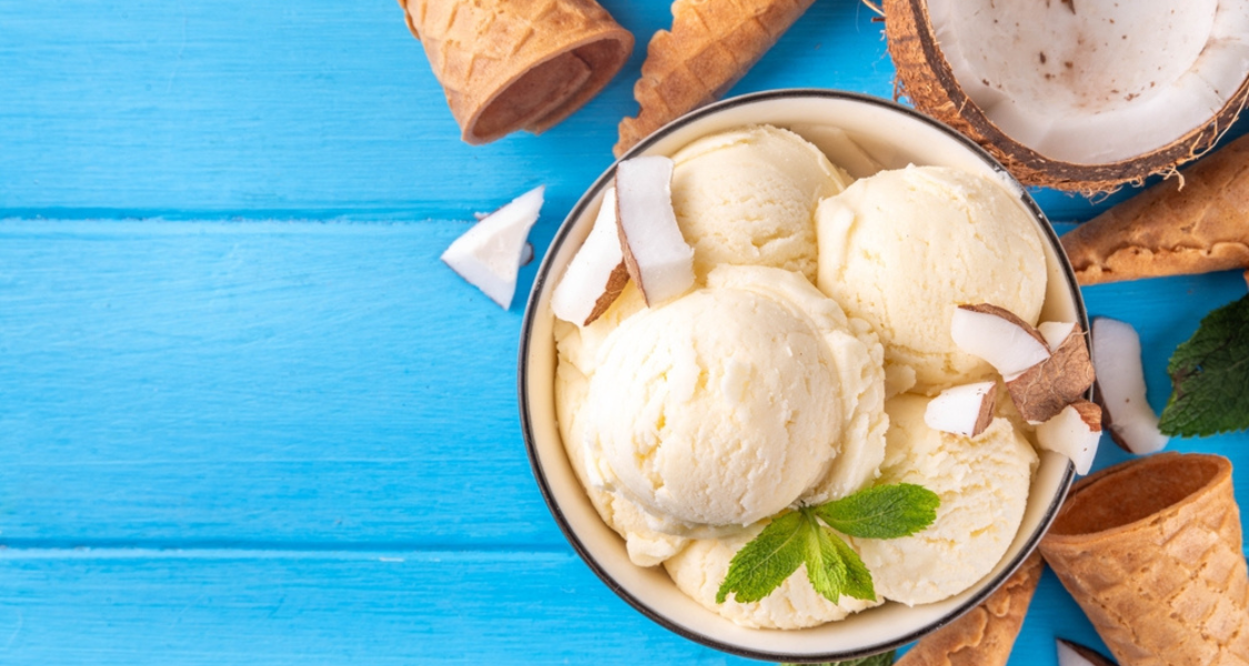 A imagem mostra uma taça de sorvete de coco vegano, com bolas cremosas de cor branca, decoradas com lascas de coco natural e uma folhinha de hortelã. Ao redor, há casquinhas de sorvete e pedaços de coco sobre uma superfície azul vibrante, transmitindo frescor e leveza.