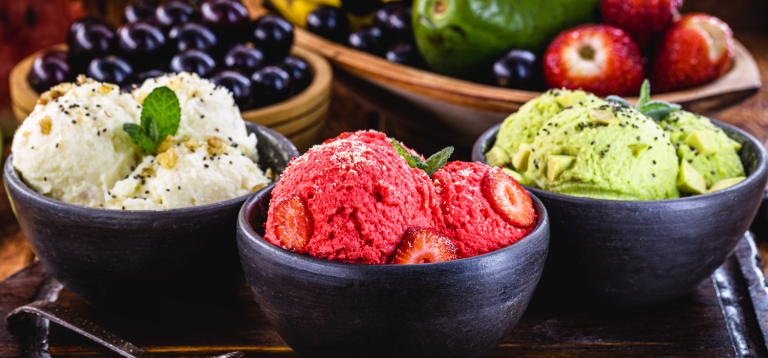 A imagem mostra três tigelas com sorvetes veganos coloridos e visualmente apetitosos: um branco com nozes e sementes, outro vermelho com morangos frescos, e um verde com pedaços de abacate e sementes. Ao fundo, há frutas frescas como uvas, morangos e abacates, reforçando a ideia de ingredientes naturais e saudáveis.