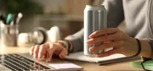 A imagem mostra uma pessoa utilizando um laptop enquanto segura uma lata de bebida, aparentemente energética, sem rótulo visível. O ambiente sugere um espaço de trabalho ou estudo, com itens como cadernos, fones de ouvido e materiais de escritório ao redor.