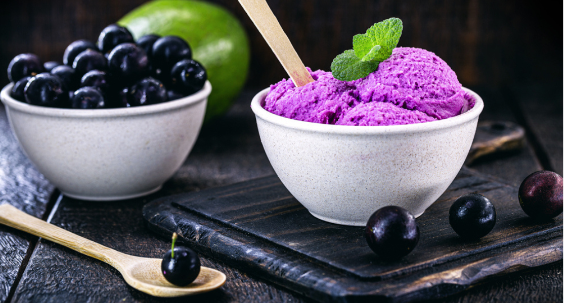 A imagem exibe uma taça de sorvete vegano roxo vibrante (possivelmente de açaí ou jabuticaba), decorado com uma folhinha de hortelã, servido em um recipiente branco rústico sobre uma tábua escura de madeira. Ao lado, há uma tigela com frutas roxas frescas e uma colher de madeira, em um ambiente que mistura rusticidade com apelo natural.