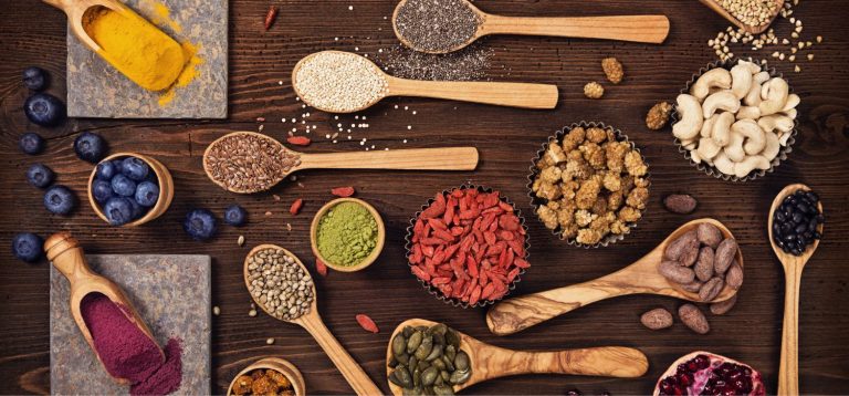 Diversas especiarias, sementes e ingredientes naturais dispostos em colher e recipientes de madeira sobre uma superfície de madeira escura. A imagem retrata aplicações dos superalimentos na indústria.