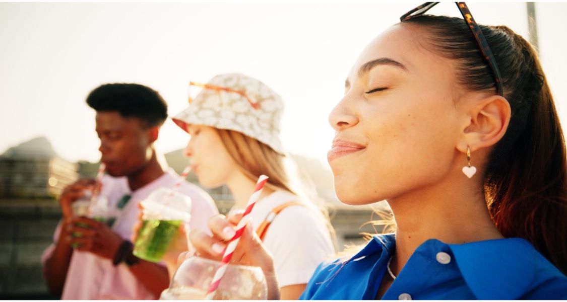 A imagem mostra três jovens ao ar livre, aproveitando um momento de lazer enquanto bebem bebidas à base de plantas em copos transparentes com canudos. A atmosfera é descontraída, iluminada por luz natural quente, sugerindo um final de tarde ensolarado.