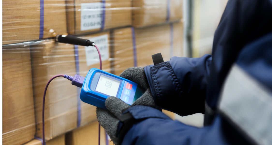 Um trabalhador com luvas e jaqueta térmica está segurando um termômetro digital conectado por fios a uma sonda inserida em uma caixa envolvida por filme plástico. O visor do aparelho exibe a leitura de temperatura interna (–6,8 °C), indicando que a carga está sob condições refrigeradas ou congeladas.