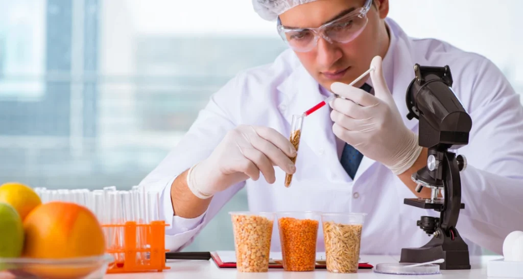 Cientista realizando análise em laboratório com microscópio, tubos de ensaio e frutas ao fundo.