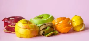 Frutas de casca e polpa coloridas, incluindo maçã, kiwi, laranja, limão e mamão, dispostas sobre fundo rosa claro, ilustrando o tema das collabs de upcycling.