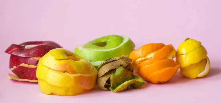 Frutas de casca e polpa coloridas, incluindo maçã, kiwi, laranja, limão e mamão, dispostas sobre fundo rosa claro, ilustrando o tema das collabs de upcycling.