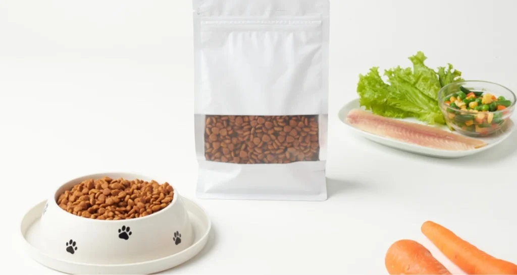 Imagem de comida para cachorro com ração, tigela, alimento natural, legumes e peixe em ambiente branco.