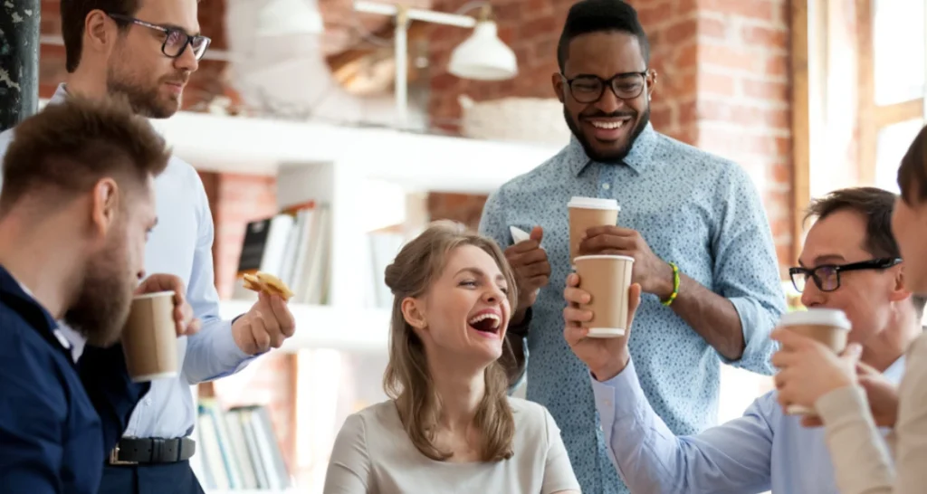 Grupo de jovens profissionais sorrindo e conversando em ambiente de escritório moderno, com alguns segurando copos de café.