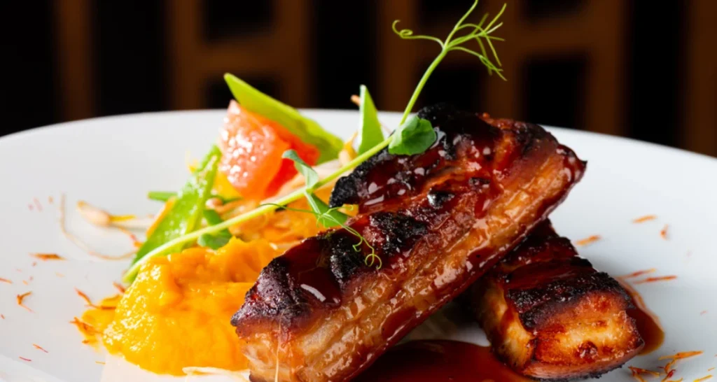 Imagem de uma deliciosa costela de porco assada com molho barbecue, acompanhada de purê de abóbora, salada de cenoura e abacate, servida em um prato branco.