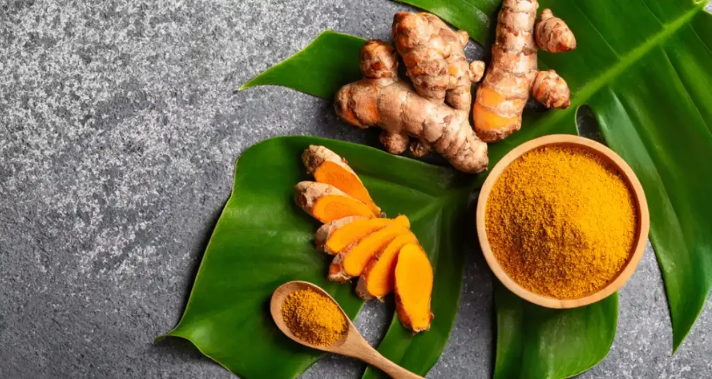 Imagem de cúrcuma fresca e em pó sobre folhas verdes, ideal para benefícios à saúde e culinária saudável.