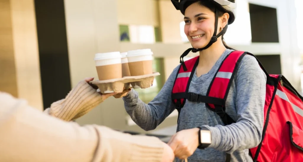 Mulher entregando café para um entregador sorridente usando capacete de bicicleta em ambiente urbano.