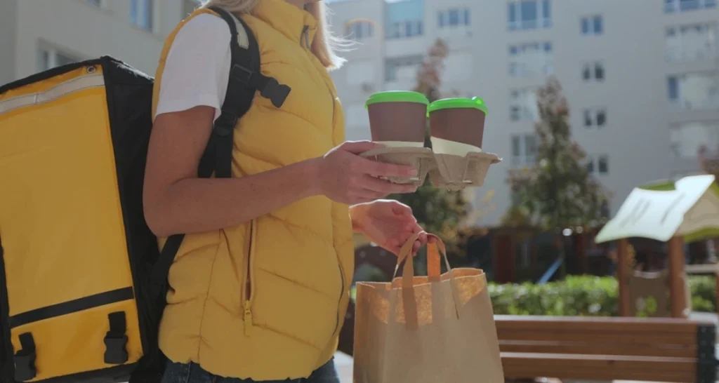 Entregadora de comida delivery com mochila amarela entregando comida em sacola de papel ao ar livre, em área urbana com prédios ao fundo.
