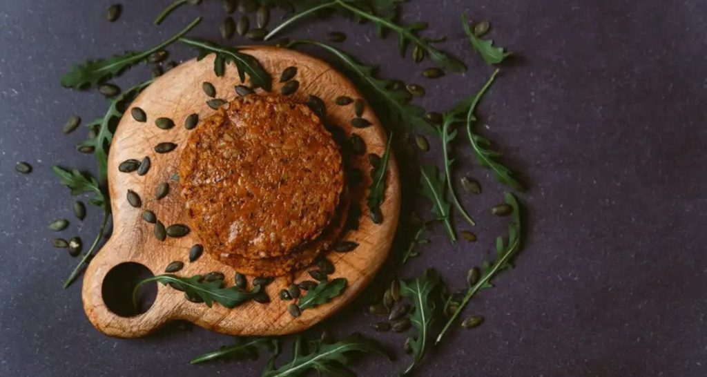 No centro da imagem, empilhados sobre uma pequena tábua de madeira rústica (com alça circular), há pelo menos dois hambúrgueres de aparência vegetal. Eles têm uma textura granulada e aparência marrom-avermelhada, indicando ingredientes como leguminosas, grãos ou vegetais como beterraba ou cenoura. Também é possível que contenham sementes ou grãos inteiros.
