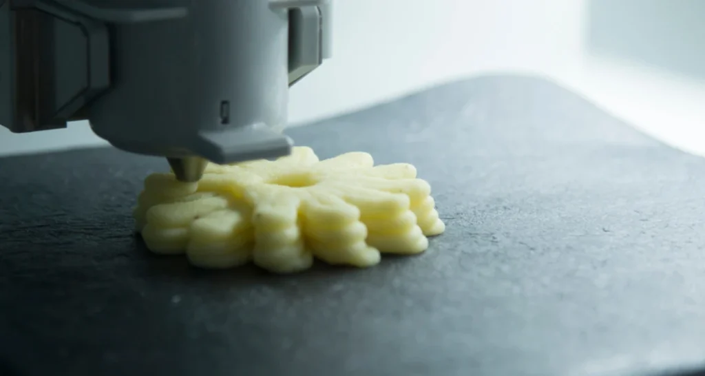 Impressora 3D criando um alimento impresso em uma superfície cinza.