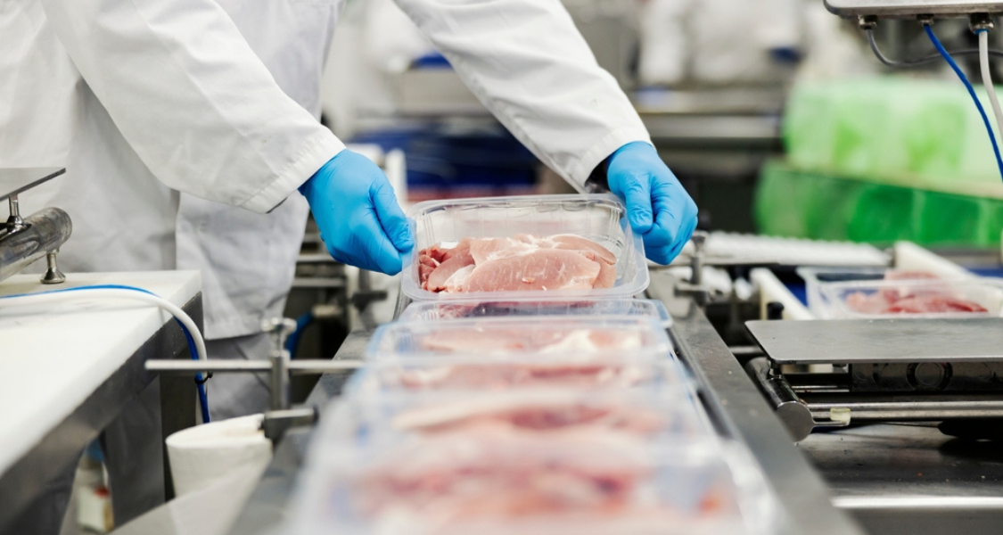 A imagem mostra um profissional em ambiente industrial manipulando bandejas de carne fresca ou resfriada, com foco em práticas de embalagem e processamento na indústria de alimentos. A seguir está uma descrição detalhada com base em logística de carnes congeladas