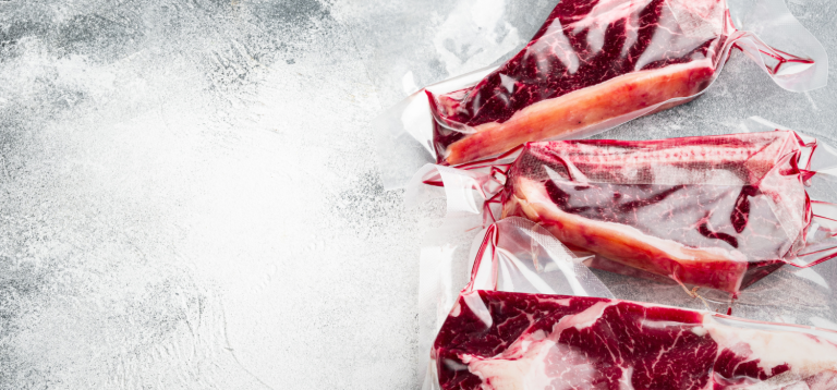 A imagem mostra cortes de carne vermelha embalados a vácuo, dispostos sobre uma superfície fria e com aparência congelada. Essa apresentação está diretamente relacionada ao armazenamento adequado de carnes congeladas.