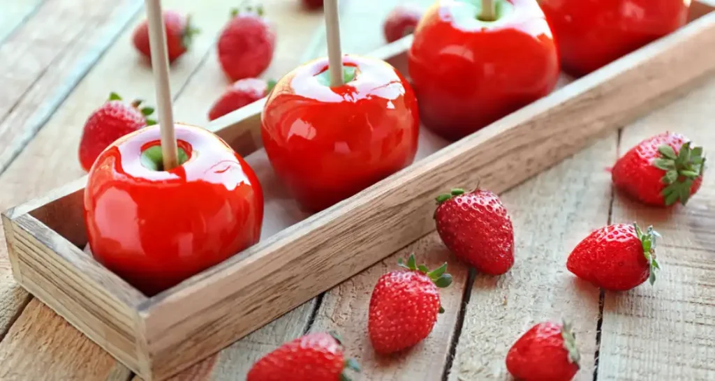 Maçãs do amor cobertas com calda vermelha e decoradas com morangos ao redor, dispostas em uma caixa de madeira sobre uma mesa rústica.