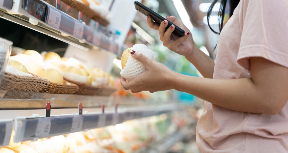 A imagem mostra uma mulher em um supermercado, segurando uma fruta protegida com malha de espuma e utilizando um celular, possivelmente para escanear um código, comparar preços ou buscar informações sobre o produto.