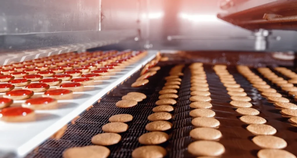 Processo de produção de biscoitos em uma fábrica, com várias formas e tamanhos de biscoitos sendo assados em uma linha de produção industrial.