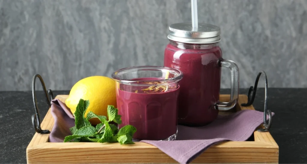 Bebida de suco de açaí em um copo de vidro, acompanhada de limão, hortelã e uma jarra de vidro com tampa, tudo disposto em uma bandeja de madeira sobre uma mesa.