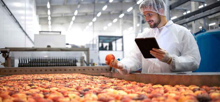 Homem em uma fábrica de alimentos, segurando uma maçã e usando um tablet, com várias maçãs ao seu redor, símbolo de controle de qualidade em produção de alimentos.