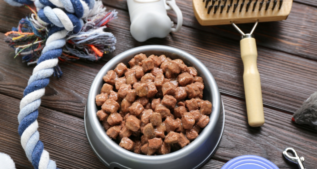 Imagem de um pote de comida para gatos com cubos de ração, ao lado de brinquedos e acessórios para gatos em uma superfície de madeira.