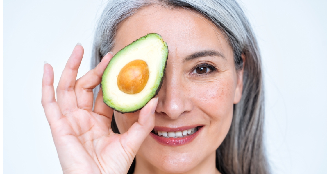 A imagem mostra uma mulher madura sorridente segurando metade de um abacate à frente de um dos olhos, como se fosse uma lente ou "máscara divertida". O fundo é branco, o que destaca ainda mais o rosto da mulher e o alimento.