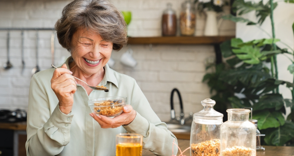 A imagem mostra uma mulher idosa sorridente, sentada à mesa em uma cozinha bem iluminada e decorada com plantas. Ela está segurando uma tigela de vidro com granola (ou cereais integrais) e usando uma colher para comer, claramente desfrutando da refeição.